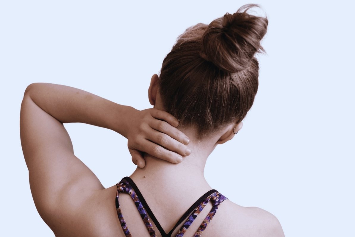 A woman seen from behind with her hand on the back of her neck, wearing a patterned athletic bra against a light blue background.